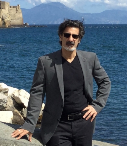 Richard Barbiero, a light-skinned person with medium-short dark hair, moustache, and graying beard wearing a black V-neck shirt under a gray suit coat, dark slacks, belt, and sunglasses, posing by a rocky escarpment on a body of water