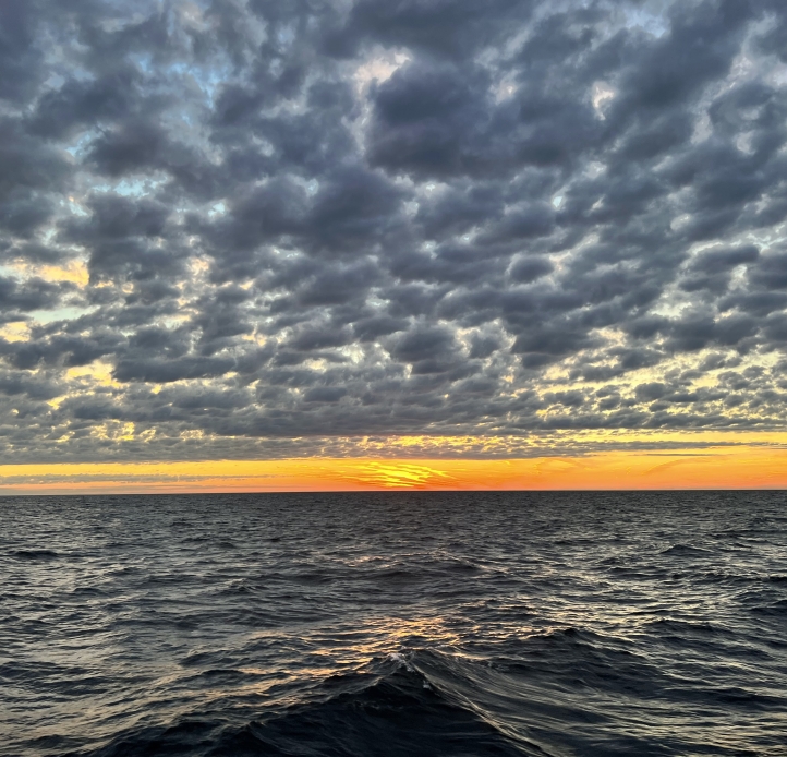 Sunset on a large body of water. A thin band of orange rests between gray dappled clouds and steely waves.