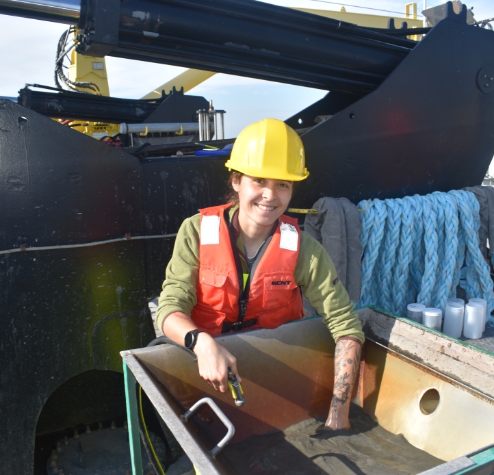 A person in a life jacket and hard hat reaches into a basin of muddy water on the deck of a ship in front of a large hydraulic winch