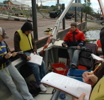 Students sit on a boat tied up at dock while a lecturer holds up a research instrument