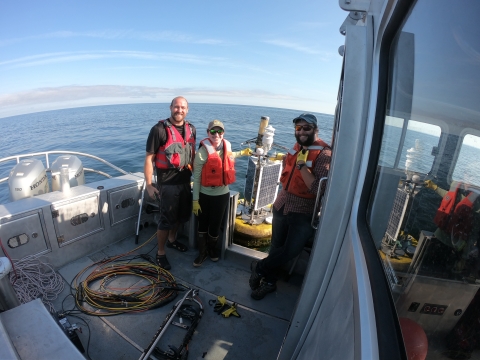 Three people smile on the back of a boat holding a squat buoy with solar panels and weather instruments.