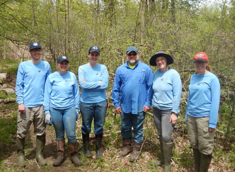 Six people posing for a picture in the woods. All wear matching work shirts, hats, and work boots.