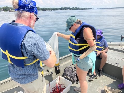 Several people on a small boat. A person with a prosthetic arm holds up a conical plankton net to drain it after collecting a sample