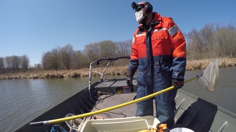 A person wearing a safety suit stands on the deck of a small jon boat. They are holding a net on a long pole and wearing a cloth mask. There is a camera strapped to their head.