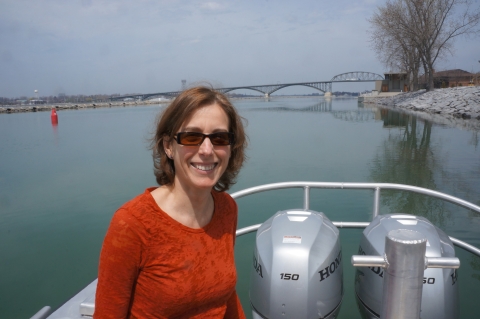 A smiling woman with sunglasses and shoulder length hair poses for a picture on the back of a boat in a canal, a large bridge in the distance