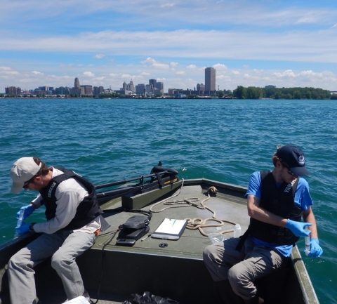 Two people collect water samples on either side of the front of a small boat. A city skyline is at the edge of the lake in the distance.