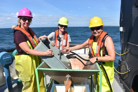 Three people wearing safety gear work on the back deck of a large boat. Two hold a tub of sediment in a concave basin table while the third sprays the tub with a hose.