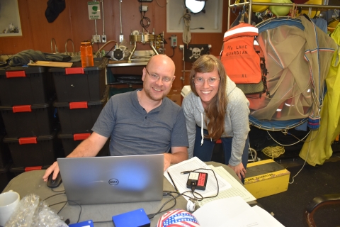 Two people pose for a picture in a workshop. One is sitting in front of a laptop at a table while the other stands next to them. There are nets, plastic totes, and life jackets behind them.