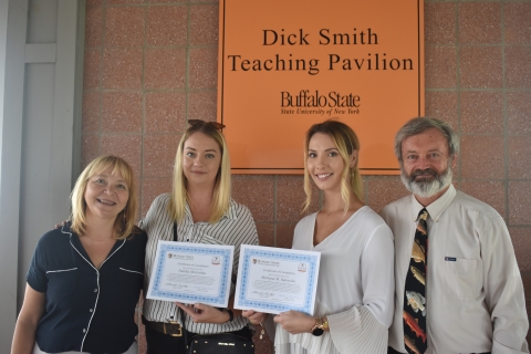 Four people posing for a picture by a wall. The two in the center are holding certificates. A sign behind them says "Dick Smith Teach Pavilion, Buffalo State."