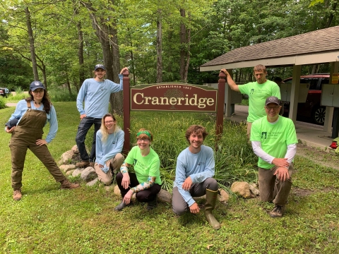 Seven people pose for a picture around a sign "Craneridge, established 1967." There are trees behind them and a shelter with mailboxes nearby.