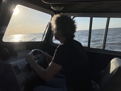 A person sits at the helm inside the cabin of a boat. The sun shines brightly through the window, casting everything inside into shade