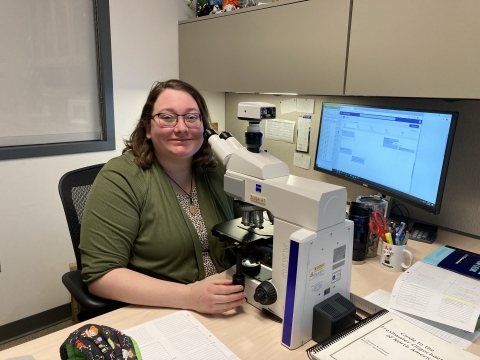 A person sits in an office in front of a microscope on her desk. She is smiling at the camera.