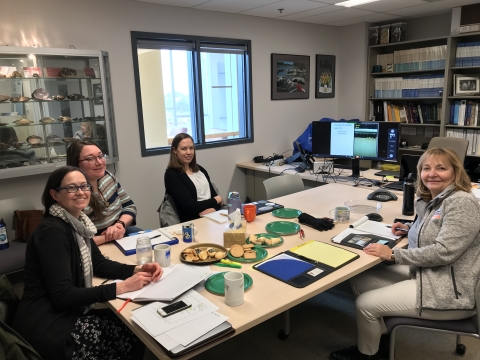 Four people sit around a table in a conference room. There are snacks in the center of the table, and everyone has paper or notebooks in front of them.