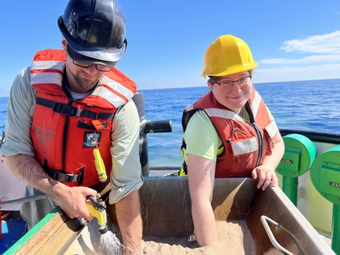 Two people wearing work gear on a boat in a large body of water reach their hands into a basin of muddy water that one is filling with a hose.