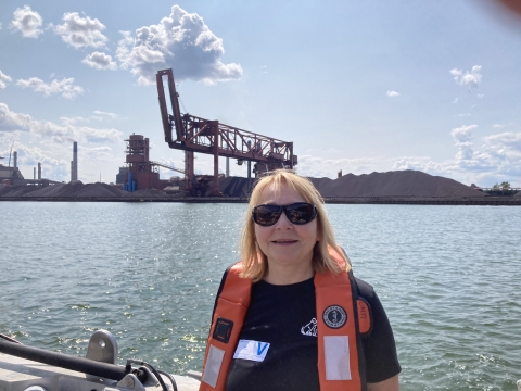 A person in sunglasses poses in front of a body of water with industrial cranes and piles of stones