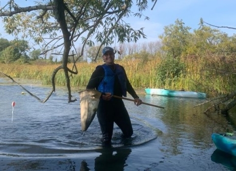 A person wading in shallow water pauses to smile for the camera. They're wearing a wetsuit and life jacket and holding a net on a pole. There is a kayak parked by some tall wetland plants at the edge of the water.