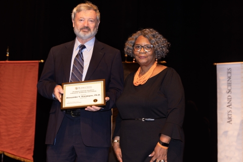 Two people pose on stage while one holds a framed award