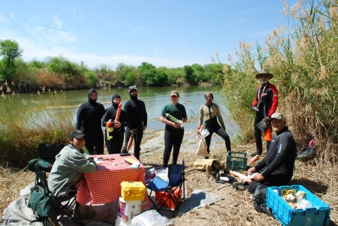 Eight people in wet suit posing for a picture by a river. They have sampling gear and a table for working on.