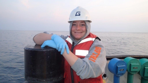 A portrait of a person leaning on a post at the edge of a boat. They are wearing a hoodie with the hood up under a hard hat, rubber gloves, and a life jacket.