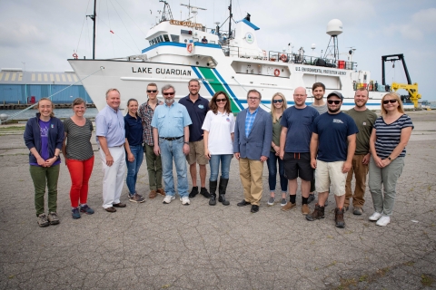 Fifteen people pose for a picture outside. There is a large boat behind them labeled "Lake Guardian."