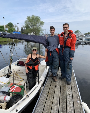 Two people stand on a dock near a boat with a top while another person stands in the boat next to them. They are all wearing full body flotation suits.