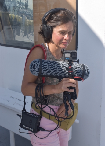 A person holding a Go Pro camera and a large horizontal microphone while wearing headphones and recording equipment