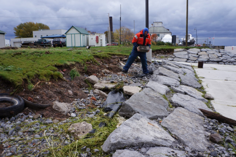 A person stands in a debris-filled area between neatly laid stone and eroded grass and dirt.