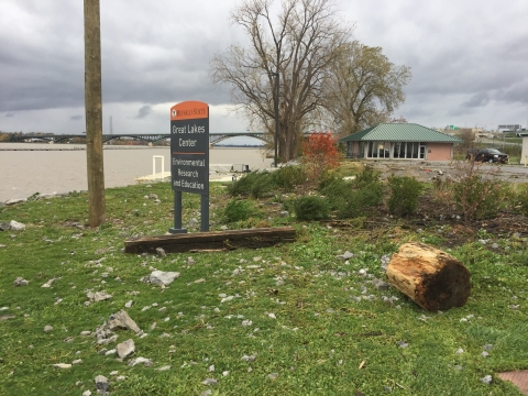 A lawn near the waterfront with a garden and a sign reading "Buffalo State Great Lakes Center." There are large pieces of wood and partial tree trunks on the lawn many yards from the water's edge, and the lawn is peppered with large stones and gravel.