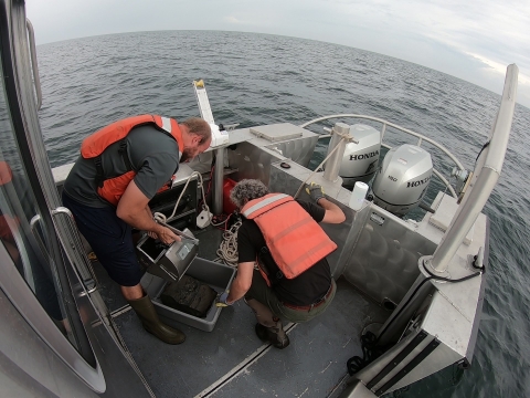 Two people work on the back deck of a boat on a body of water. One holds a metal dredge scoop above a rectangular bucket with a scoop of sediment in it while the other examines the sediment. The horizon is curved due to a fish-eye lens on the camera.