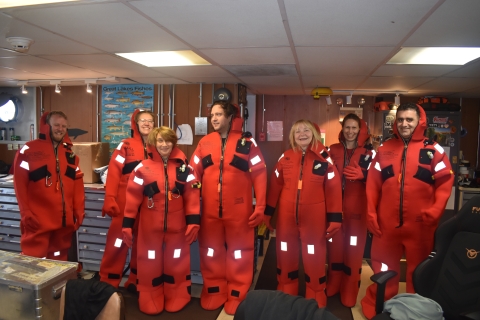 A group of seven people pose for a picture while wearing immersion suits during a training inside a boat