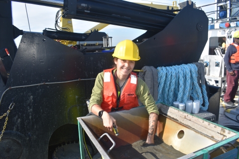 A person in a life jacket and hard hat reaches into a basin of muddy water on the deck of a ship in front of a large hydraulic winch