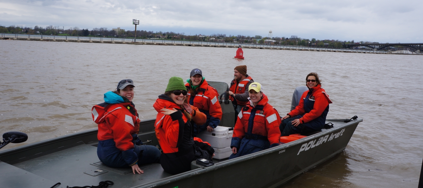 Six people in coldwater safety gear on a small boat in a muddy river on a cold day. Most are smiling or waving at the camera while one drives