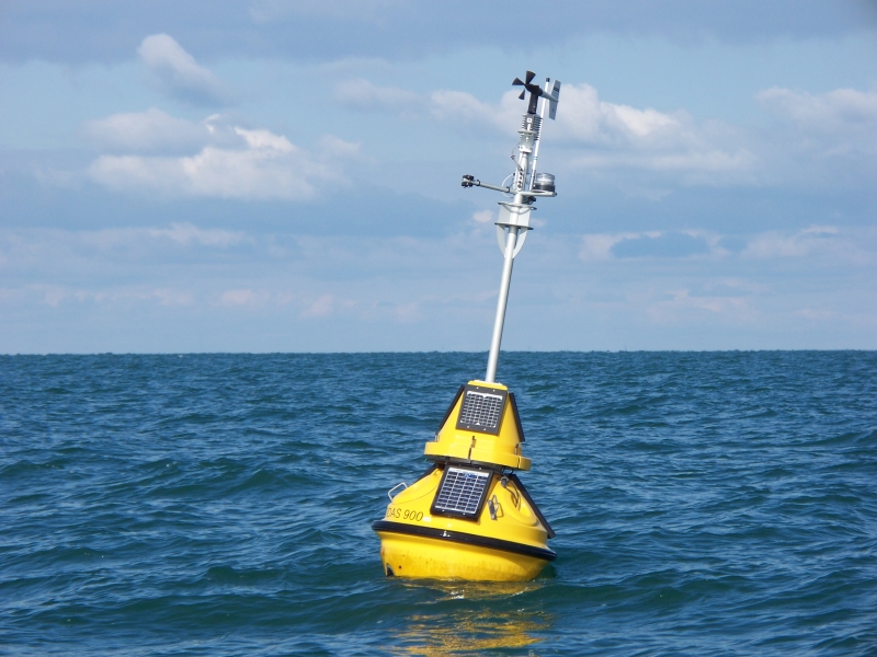 A buoy with a mast with weather instruments rocks in the water on a partly cloudy day with small waves