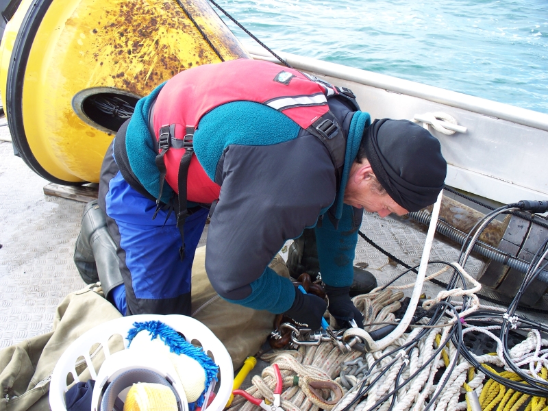 A person uses pliers to unhook two pieces of rope on the deck of a boat. A buoy is lying on the deck nearby.