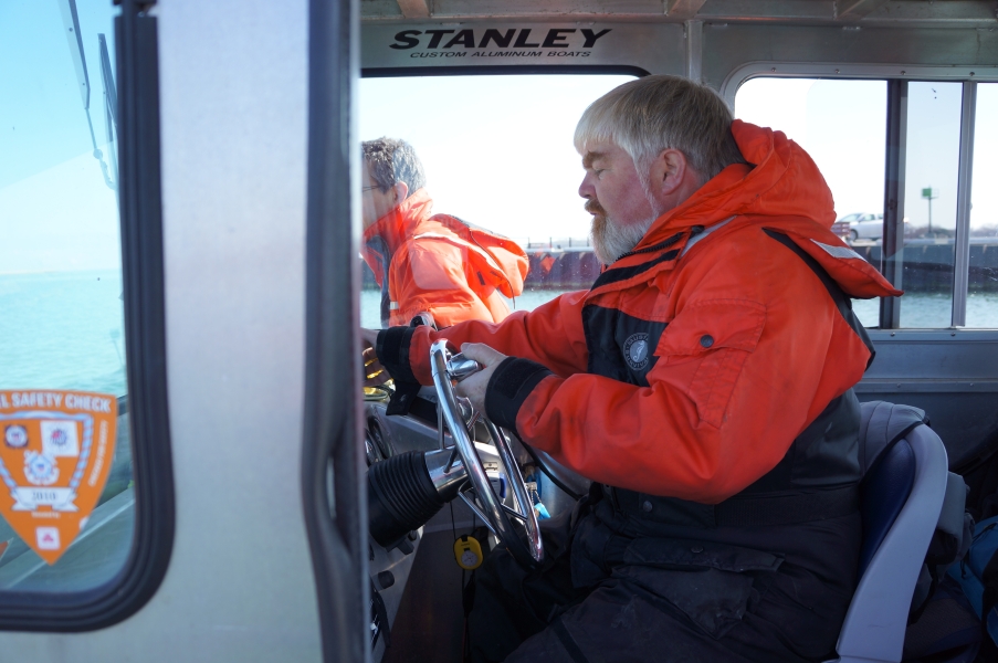 A person in a cold water safety suit sitting at the helm of a boat, while another person stands outside on the other side of the cabin