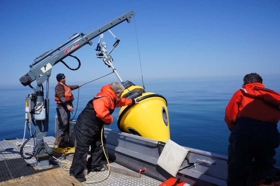 People working on the deck of a boat. There is a buoy next to the side of the boat attached to a crane that is being lowered into the water.