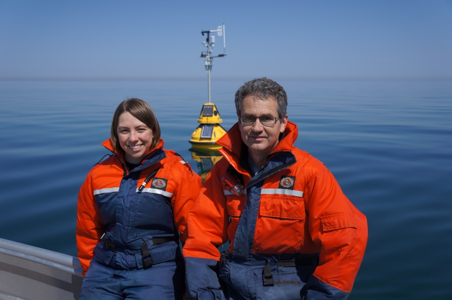 Two people in cold water safety suits sit on the edge of a boat in front of a buoy in the water.