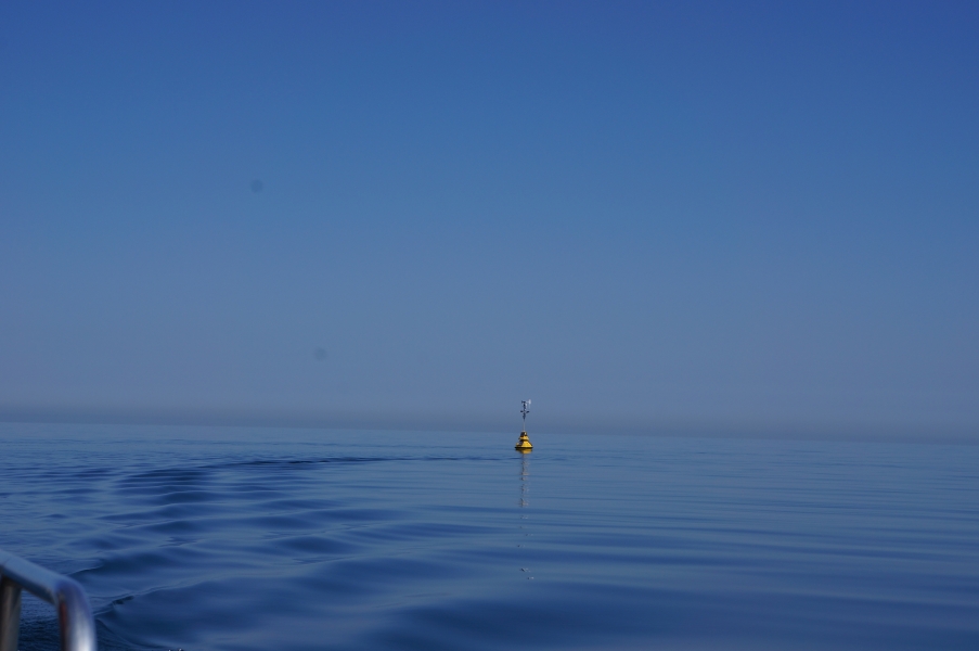 A buoy sits in calm water near the wake of a boat. There is not much distinction between the sky and the water.