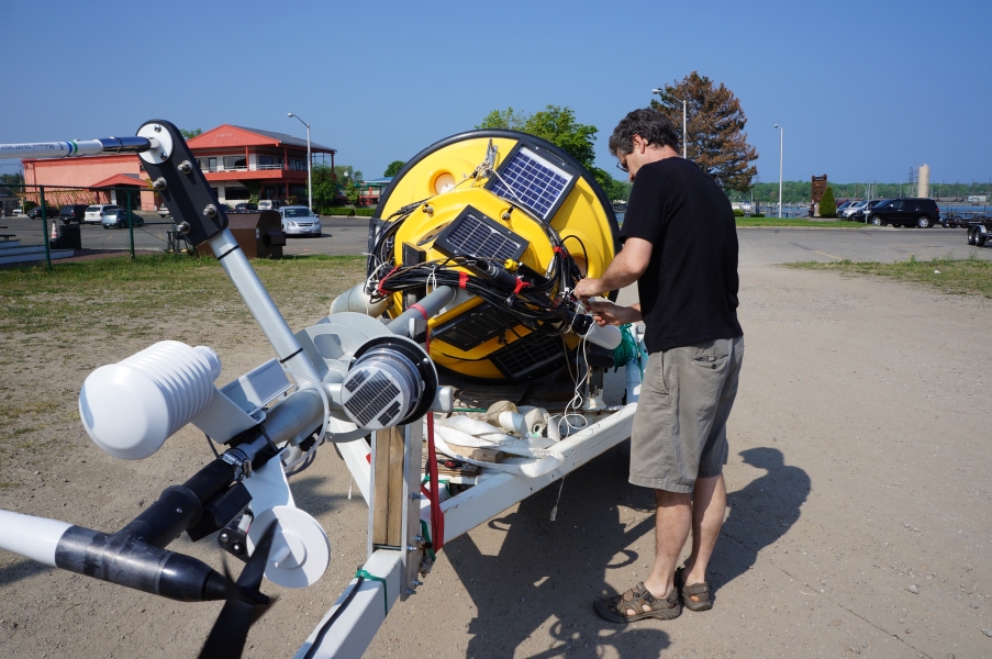 A person works on a buoy with weather instruments on a mast sitting on a boat trailer in a parking lot