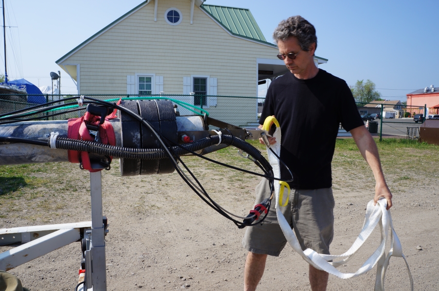 A person loops a white strap through the bail at the end of buoy that's lying on a trailer. There are heavy weights and cables at this end of the buoy.