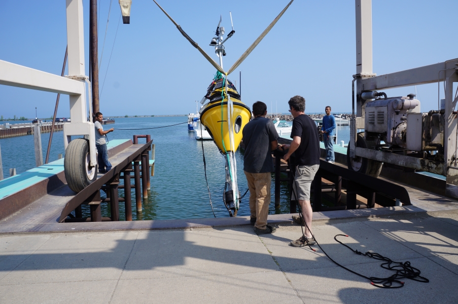 A buoy hangs over the water from a strap on a large metal frame. Two people are guiding the bottom end of the buoy out over the water