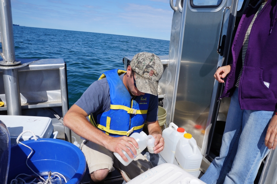 A person on a boat pours something from one jar into a smaller bottle. A second person stands by, and there are jugs and coolers on the back of the boat with them.