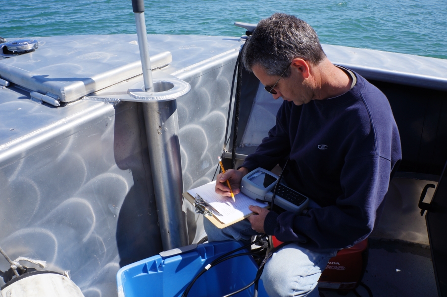 A person on a boat holds an instrument in their lap while they write something on a clipboard