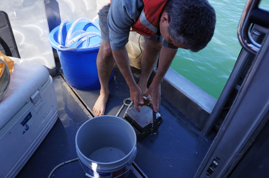 A person on a boat holds a metal grab sampler on the deck of a boat.