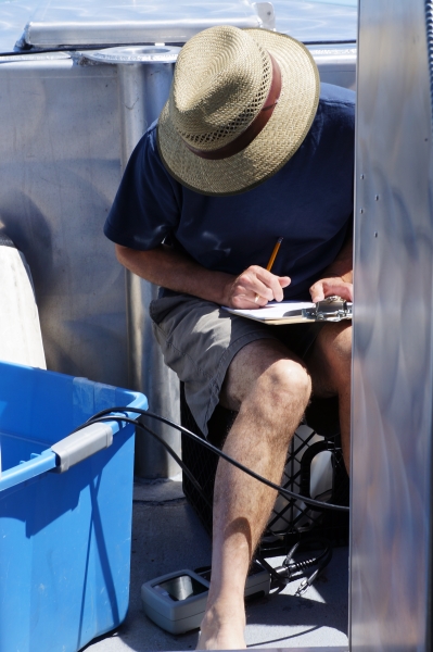 A person with a hat obscuring their face works on a boat. They are sitting on a milk crate, looking down at an instrument on the floor, and writing something on a clipboard.