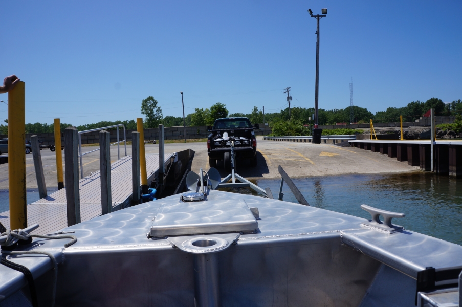 The bow of a boat near a dock. A truck with a trailer is slightly ahead of the boat near the edge of the water.