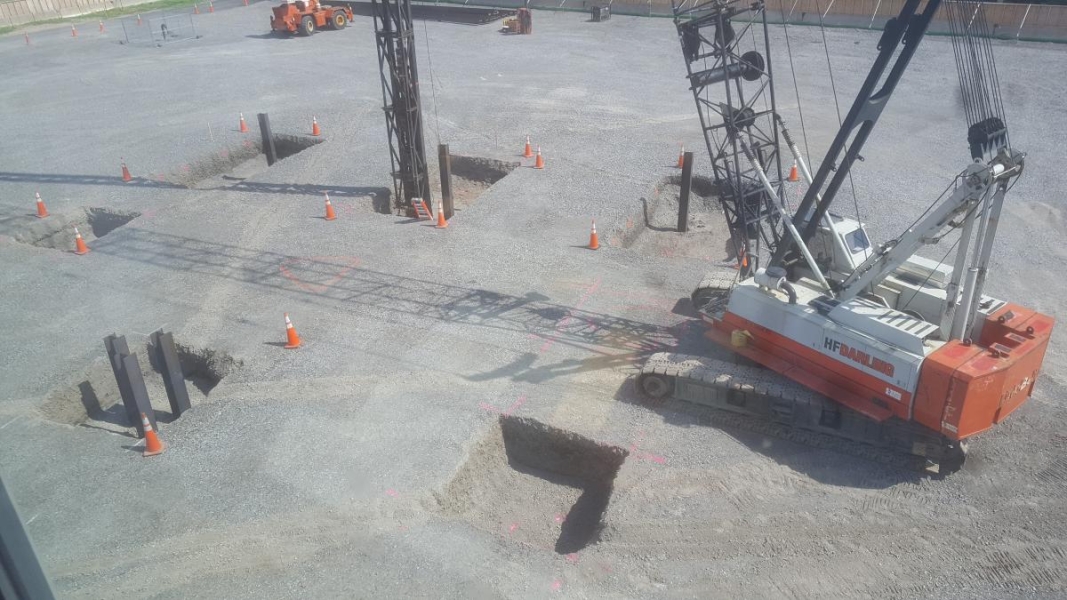 A crane pounding steel beams into sqaure holes in the ground. There are six holes and each have multiple beams driven into them.