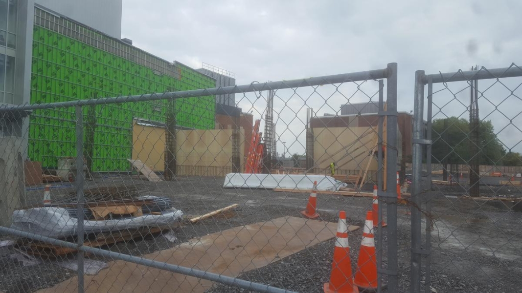 A construction site through a chain-link fence. The siding has been pulled off an existing building and the forms of new walls and rebar columns rise out of the ground next to it.