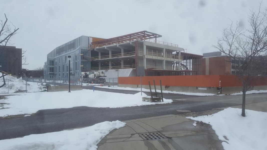 A partially constructed building. The constructed part of the building is steel and glass. The part being built is partially concrete and partially steel. The concrete part has steel beams on the top level.