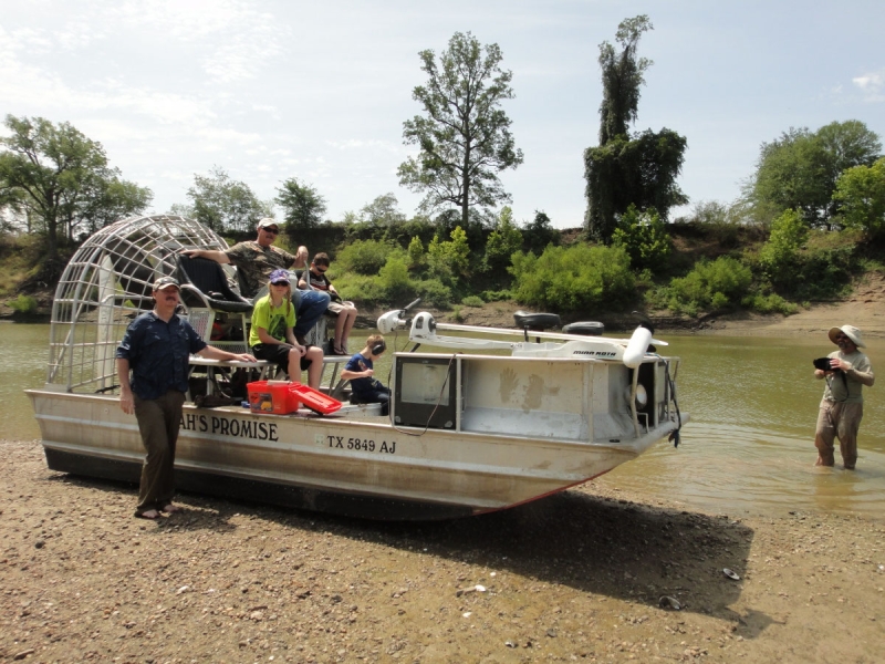 A boat with a large fan on the back sits on the sand. There are a few people on the boat, including children. One person stands on land next to the boat, and one person stands in the water.